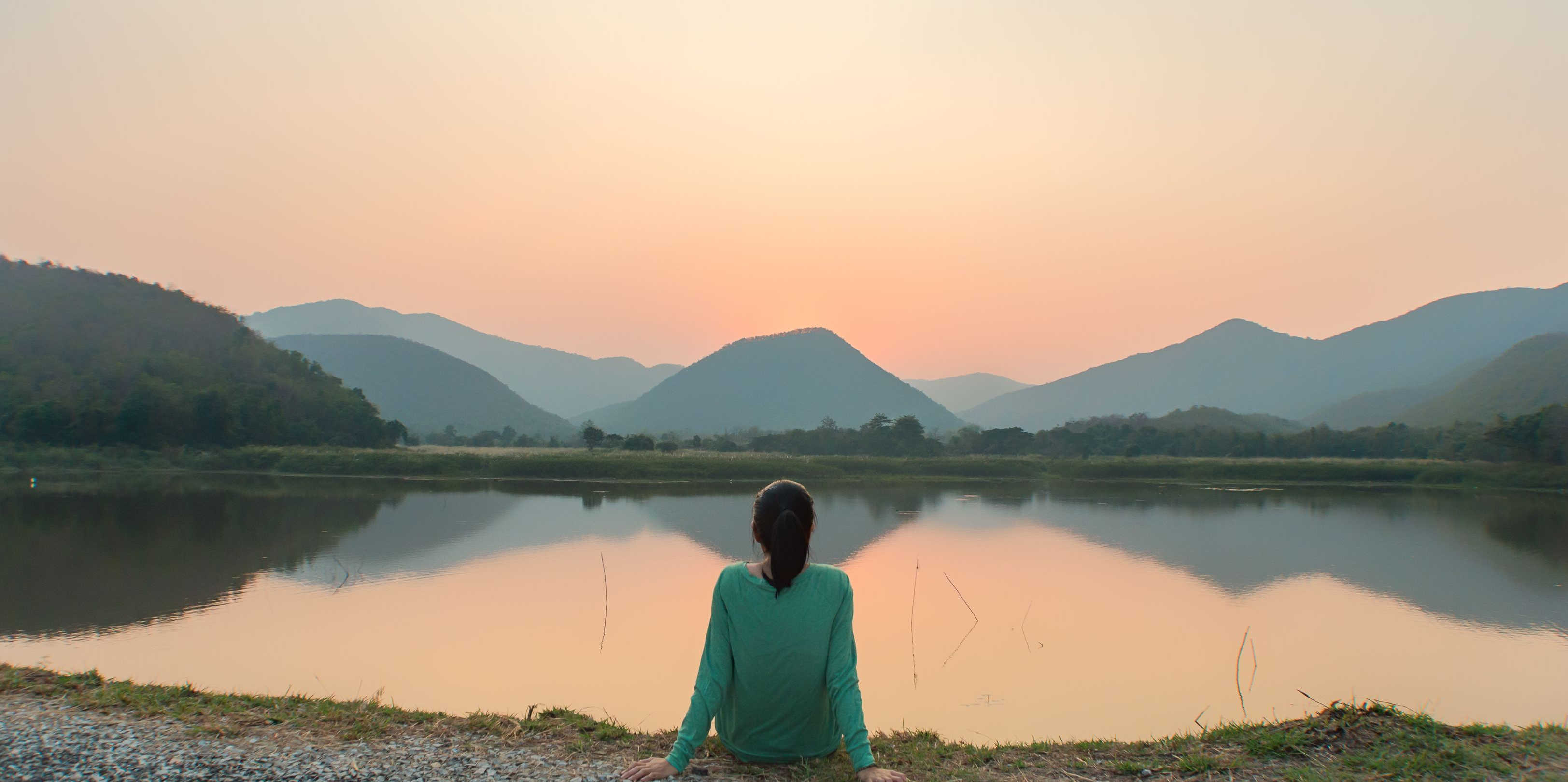 Woman looking looking at a sunset over a still lake with mountains in the background