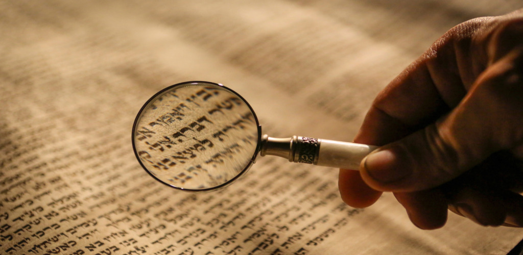 Hand holding a magnifying glass over a Hebrew manuscript